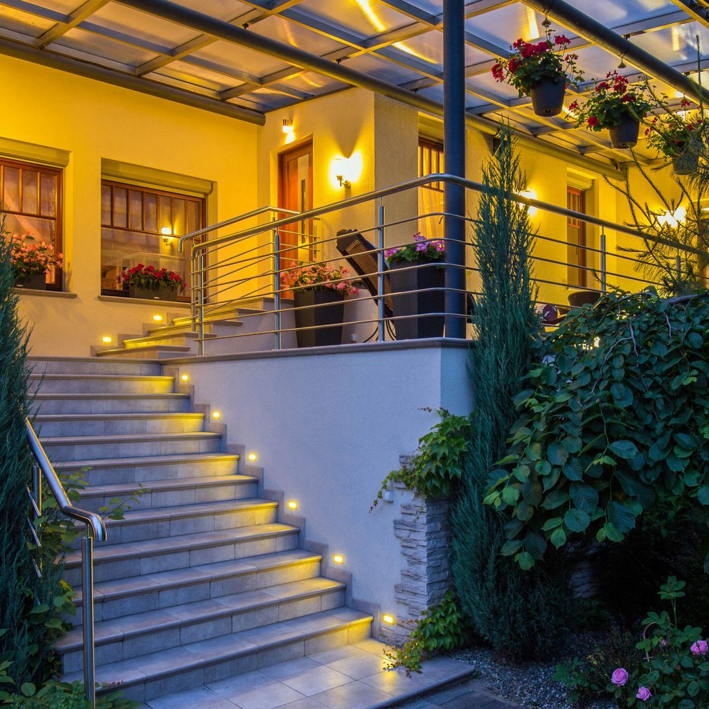 alt="Escalera y jardín iluminados con luz amarilla decorativa proveniente de bombillas LED MATEL instaladas en el techo exterior"